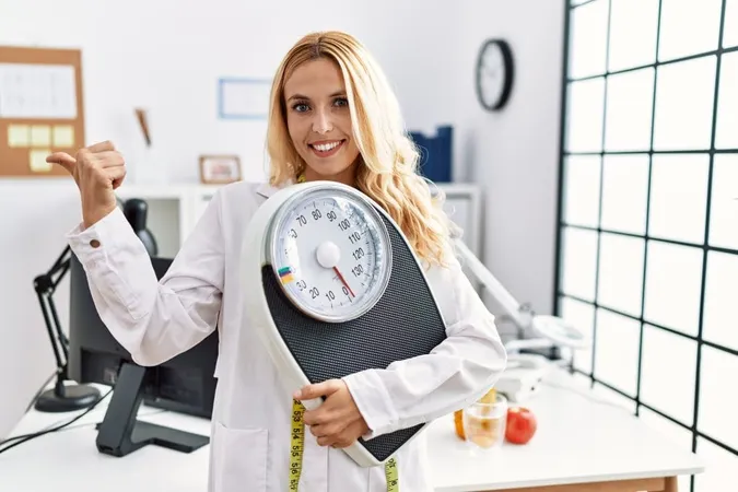 Beautiful Blonde Nutritionist Woman Holding Weight Machine Balance Weight Loss Pointing Thumb Up Side Smiling Happy With Open Mouth X - Certified Bariatric Educator study guide